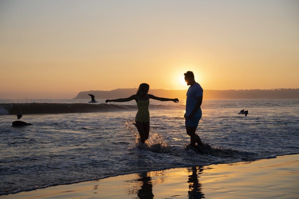 A couple enjoys walking in shallow ocean waves at sunset, with the sun setting behind them and birds flying over the sea. The sky is golden, creating a peaceful, romantic beach scene.
